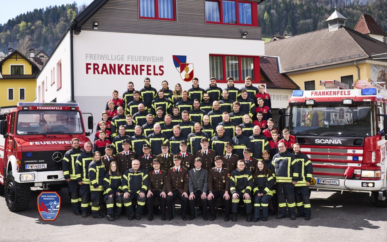 Eine Gruppe Feuerwehrleute in Uniform posiert für ein Foto vor ihrem Stützpunkt, einige stehend und andere auf einer Bank sitzend, umgeben von Feuerwehrwagen.