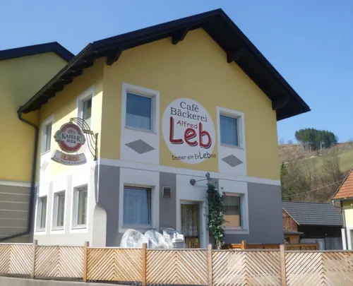 Ein gelbes Gebäude mit einem braunen Dach hat ein Schild mit der Aufschrift Café Bäckerei Alfred Leb. Das Gebäude hat einen Zaun und eine Bank davor.