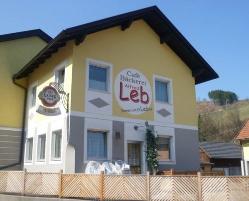 Ein gelbes Gebäude mit einem braunen Dach hat ein Schild mit der Aufschrift Café Bäckerei Alfred Leb. Das Gebäude hat einen Zaun und eine Bank davor.