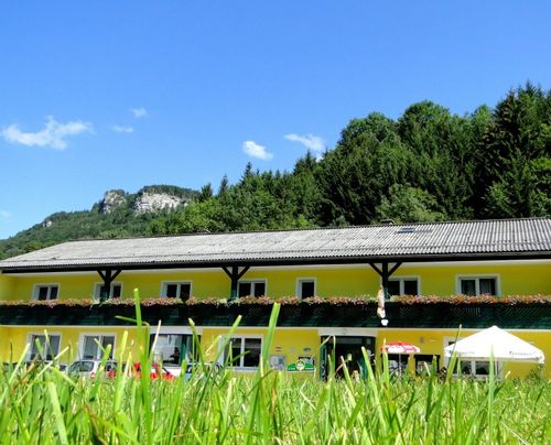 Ein strahlend gelbes Hotel mit mehreren Fenstern und einem Balkon voller Blumen steht vor einem grünen Hang unter einem blauen Himmel.
