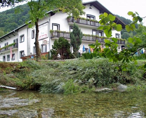 Ein weißes Gebäude namens Hotel Pension steht am Hang eines Flusses. Es ist von Bäumen und Pflanzen umgeben, mit einer Flagge links und Schirmen davor.