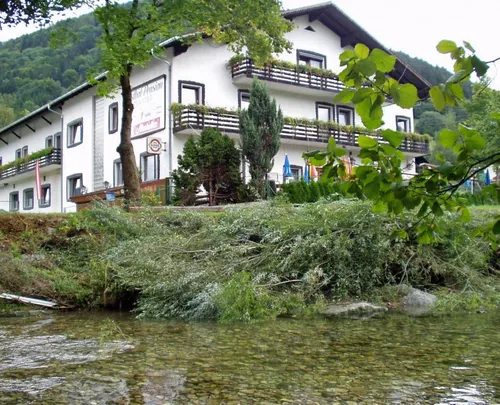 Ein weißes Gebäude namens Hotel Pension steht am Hang eines Flusses. Es ist von Bäumen und Pflanzen umgeben, mit einer Flagge links und Schirmen davor.