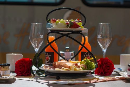 Ein schwarzer Ständer hält eine Obstplatte mit Trauben, Erdbeeren und einem Weinkelch. Darunter ist ein Teller mit Käse, Fleisch und Gemüse. Zwei rote Rosen sitzen auf beiden Seiten des Ständers.
