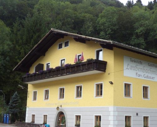 Ein gelbes Gebäude mit Balkon trägt ein Schild mit der Aufschrift Tankstelle Laubenbach Fam. Gab. Es hat Topfpflanzen auf dem Balkon und zwei rote Regenschirme.
