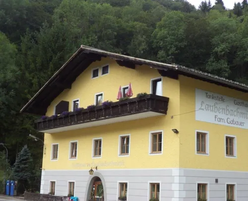 Ein gelbes Gebäude mit Balkon trägt ein Schild mit der Aufschrift Tankstelle Laubenbach Fam. Gab. Es hat Topfpflanzen auf dem Balkon und zwei rote Regenschirme.