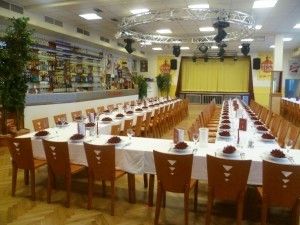 Ein Bankettsaal mit einem langen Tisch für eine formelle Veranstaltung. Der Tisch ist mit weißem Tuch bedeckt, und jede Platzierung umfasst ein Weinglas, einen Teller und eine rote Blume.