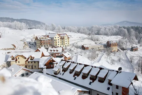 Luftaufnahme eines verschneiten Dorfes mit mehreren Gebäuden, darunter ein großes Gebäude mit Balkon und schneebedeckten Dächern, umgeben von schneebedeckten Bäumen und Bergen.