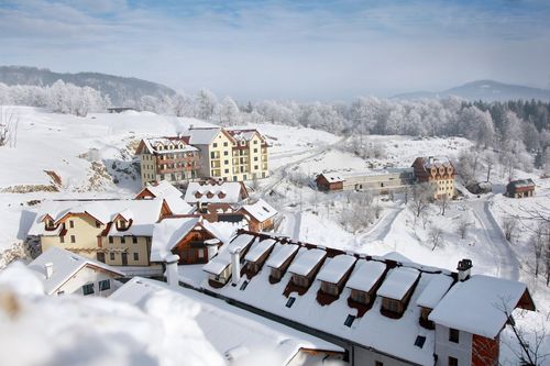 Luftaufnahme eines verschneiten Dorfes mit mehreren Gebäuden, darunter ein großes Gebäude mit Balkon und schneebedeckten Dächern, umgeben von schneebedeckten Bäumen und Bergen.