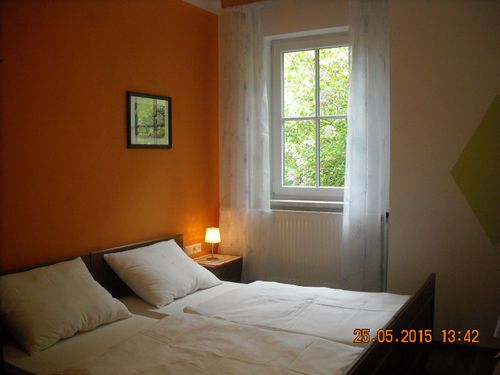 Ein Doppelbett mit weißer Bettwäsche und Kissen steht in einem Raum mit orangefarbenen Wänden. Ein Nachttisch mit Lampe steht neben dem Bett. Ein gerahmtes Bild ist über dem Bett. Ein Fenster mit weißen Vorhängen hat einen Blick auf Bäume draußen.