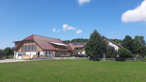 Eine ländliche Szene mit einem großen Haus mit rotem Dach und Solarpaneelen, einem Baum und einem Grasfeld. Zwei Autos sind vor dem Haus geparkt.