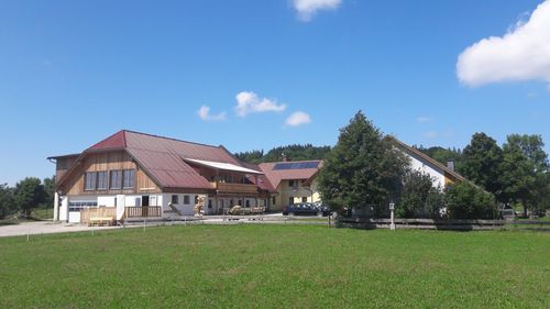 Eine ländliche Szene mit einem großen Haus mit rotem Dach und Solarpaneelen, einem Baum und einem Grasfeld. Zwei Autos sind vor dem Haus geparkt.