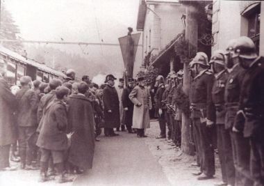 Ein Schwarz-Weiß-Foto zeigt eine Reihe von Militärangehörigen in Uniform, Helmen und Stiefeln in Formation stehend. In der Mitte unterhalten sich zwei Männer in langen Mänteln, möglicherweise bei einer Parade oder einem formellen Ereignis.