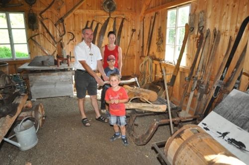 Eine vierköpfige Familie steht in einer Werkstatt mit Holzwänden und Werkzeugen. Ein Mann hält ein Stück Holz, während zwei Kinder ihn anschauen.