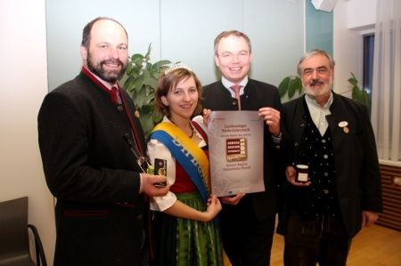 Eine Gruppe von Menschen posiert für ein Foto, während sie ein Zertifikat und eine Flasche in der Hand halten. Eine Frau in einem grünen Kleid hält ein Zertifikat. Dahinter stehen zwei Topfpflanzen und ein Glasfenster.