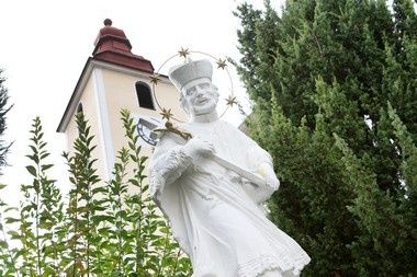 Eine Statue eines Heiligen mit Sternen steht vor einer Kirche, umgeben von üppigem Grün und einem Glockenturm mit Uhr.
