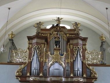 Eine große Orgel mit kunstvollen goldenen und braunen Designs ist an der Wand montiert und zeigt Statuen von Engeln oben. Sie wird durch Licht beleuchtet, das durch das Fenster kommt.