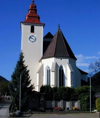 Eine weiße Kirche mit rotem Dach und Turm steht an einem sonnigen Tag. Ein Uhr ziert die Fassade des Turms.