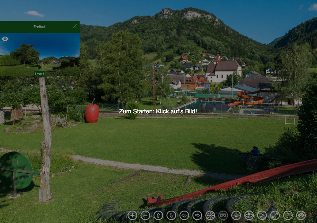 Ein Grasfeld mit einem großen roten Apfel, einem Spielplatzrutsche und einer roten Wasserrutsche. Ein Haus mit rotem Dach und einer Kirche im Hintergrund. Ein Berg mit Bäumen in der Ferne.