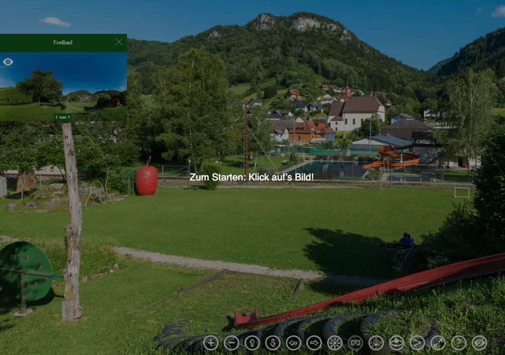 Ein Grasfeld mit einem großen roten Apfel, einem Spielplatzrutsche und einer roten Wasserrutsche. Ein Haus mit rotem Dach und einer Kirche im Hintergrund. Ein Berg mit Bäumen in der Ferne.