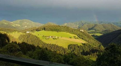 Von einem hohen Aussichtspunkt aus zeigt sich ein Tal mit grünen Feldern und Wäldern, einem Regenbogen am Himmel und fernen Bergen.