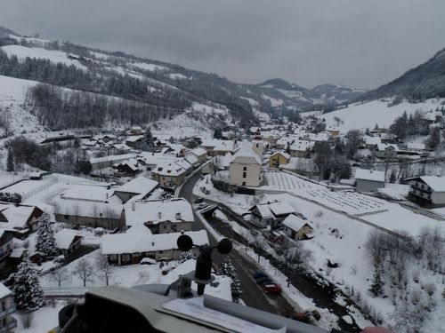 Ein verschneiter Ort in einem Gebirgsgebiet mit mehreren Häusern, einige mit schneebedeckten Dächern, einer Kirche und einer kurvenreichen Straße.