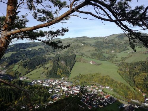 Luftaufnahme eines Dorfes, umgeben von grünen Hügeln und üppigen Wäldern, unter einem klaren blauen Himmel.