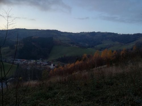 Eine hügelige Landschaft mit einem Dorf darunter und einem bewölkten Himmel. Bäume mit gelben Blättern säumen den Vordergrund, während sich grüne Felder und Hügel in die Ferne erstrecken.