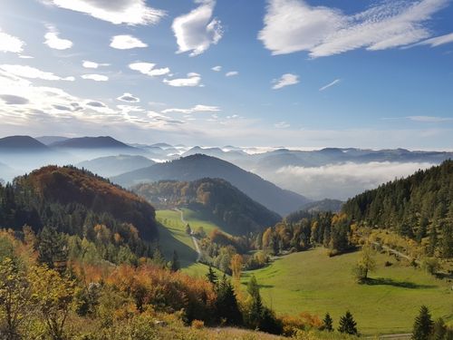 Eine bergige Landschaft mit einem friedlichen Tal, üppigen grünen Feldern und einem nebligen Horizont unter einem klaren blauen Himmel mit verstreuten Wolken.