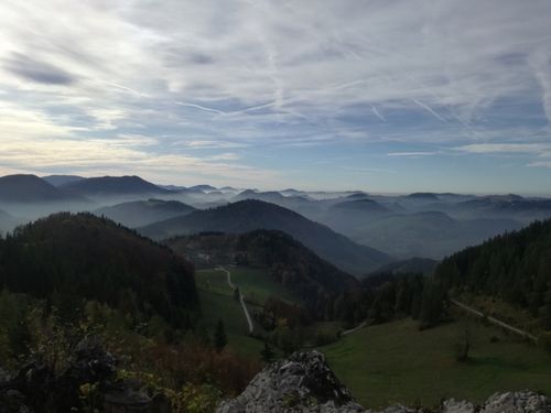 Eine Berglandschaft mit einem Tal darunter. Nebel füllt das Tal, während der Himmel teilweise bewölkt ist. Bäume bedecken die Berghänge. Ein Pfad schlängelt sich durch das Tal.