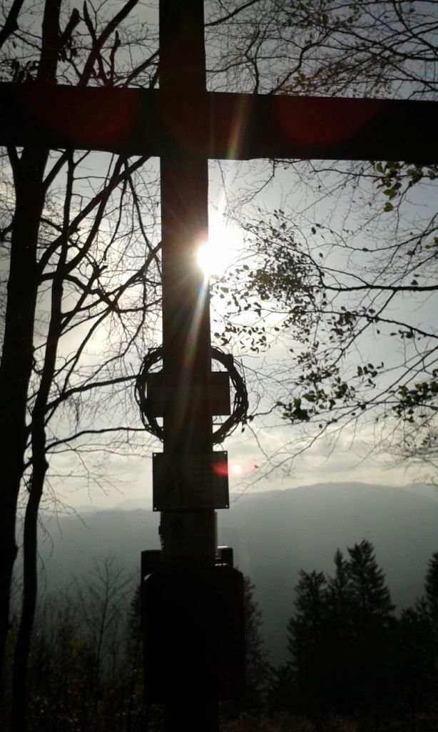Ein Kreuz steht hoch mit einem Kranz, umgeben von Bäumen. Die Sonne scheint hell dahinter und beleuchtet die Landschaft darunter.
