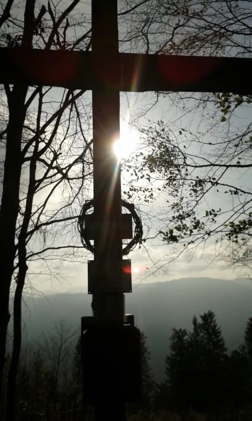 Ein Kreuz steht hoch mit einem Kranz, umgeben von Bäumen. Die Sonne scheint hell dahinter und beleuchtet die Landschaft darunter.