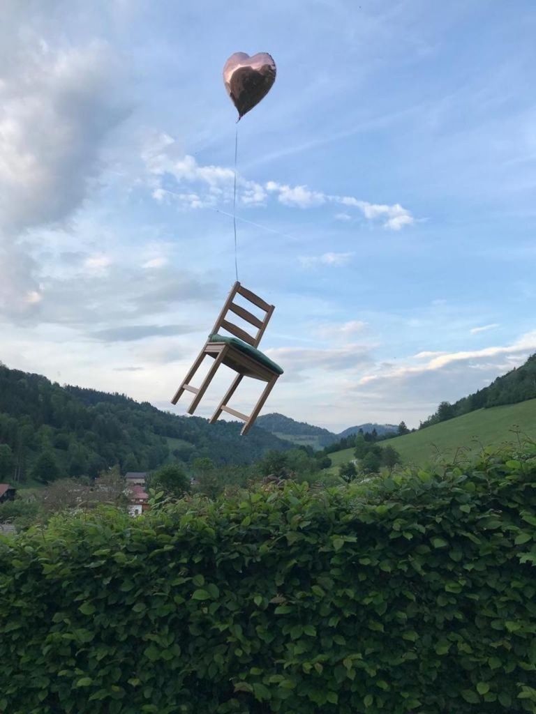 Ein Stuhl wird von einem Ballon über einer Hecke und grünen Hügeln unter einem bewölkten Himmel gehoben.