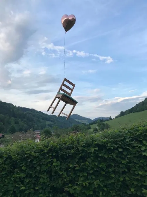 Ein Stuhl wird von einem Ballon über einer Hecke und grünen Hügeln unter einem bewölkten Himmel gehoben.