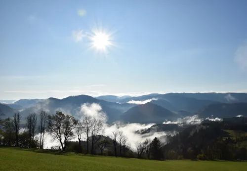 Ein strahlender Sonnenschein über einem Tal mit einem grünen Feld und kahlen Bäumen, umgeben von nebeligen Bergen.