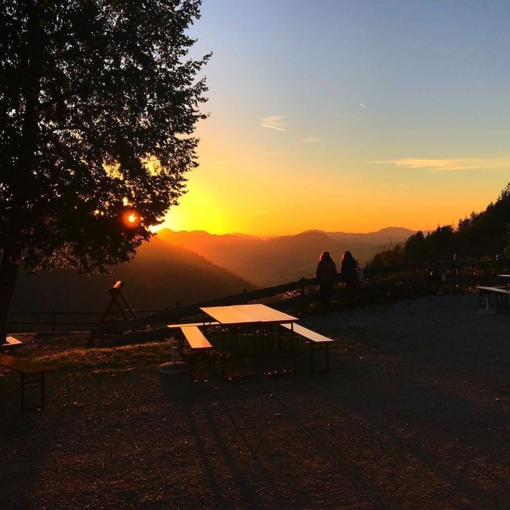 Ein Sonnenuntergang in den Bergen, bei dem ein Paar von einem Picknicktisch unter einem Baum aus zusieht. Der Himmel ist orange und pink, mit Bergen in der Ferne.