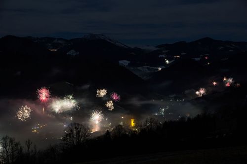 In einer dunklen Nacht mit Bergen und einer Stadt darunter sind Feuerwerke zu sehen. Ein gelbes Licht sticht in der Mitte der Stadt hervor.