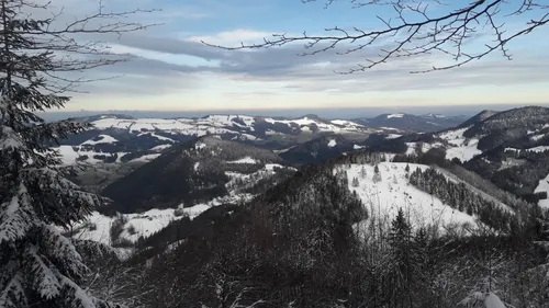 Eine verschneite Landschaft mit Bergen und Bäumen. Der Himmel ist blau mit einigen Wolken. Der Schnee bedeckt den Boden und einige Teile der Bäume.