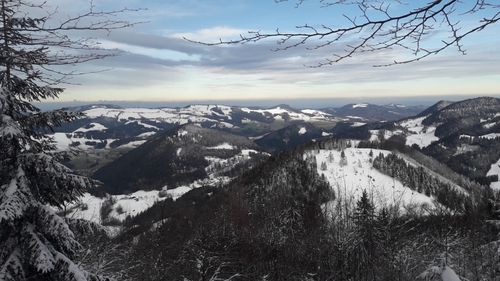 Eine verschneite Landschaft mit Bergen und Bäumen. Der Himmel ist blau mit einigen Wolken. Der Schnee bedeckt den Boden und einige Teile der Bäume.