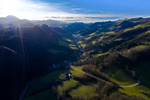 Luftaufnahme eines Tals mit üppigen grünen Feldern, verstreuten Häusern und einer kurvenreichen Straße, umgeben von dichten Wäldern und Hügeln unter einem blauen Himmel.
