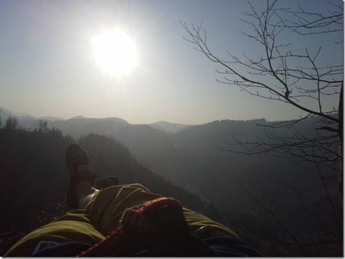 Eine Person liegt auf einem Berggipfel und blickt zum hellen Sonnenschein und zur umliegenden Landschaft hinauf.