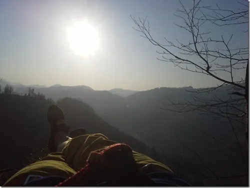 Eine Person liegt auf einem Berggipfel und blickt zum hellen Sonnenschein und zur umliegenden Landschaft hinauf.