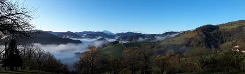 Eine neblige Berglandschaft mit grünen Hügeln, Laubbäumen und einem fernen Gipfel unter einem klaren blauen Himmel.