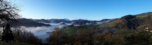 Eine neblige Berglandschaft mit grünen Hügeln, Laubbäumen und einem fernen Gipfel unter einem klaren blauen Himmel.