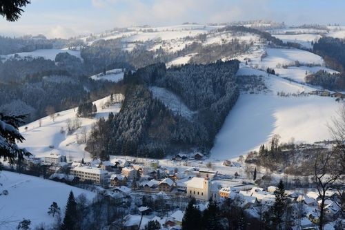 Ein verschneites Dorf umgeben von Hügeln und immergrünen Bäumen. Gebäude sind schneebedeckt, mit einer Kirche im Zentrum.