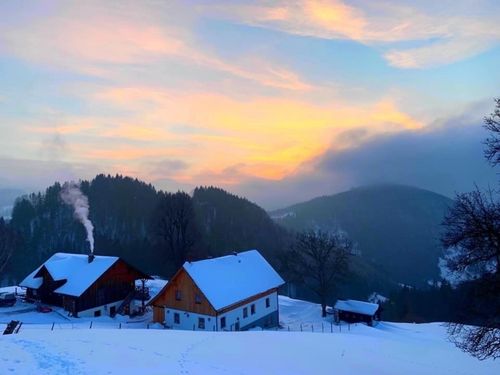 Eine verschneite Landschaft mit Häusern und einer Bergkette im Hintergrund. Der Himmel ist eine Mischung aus blauen und orangefarbenen Farbtönen.