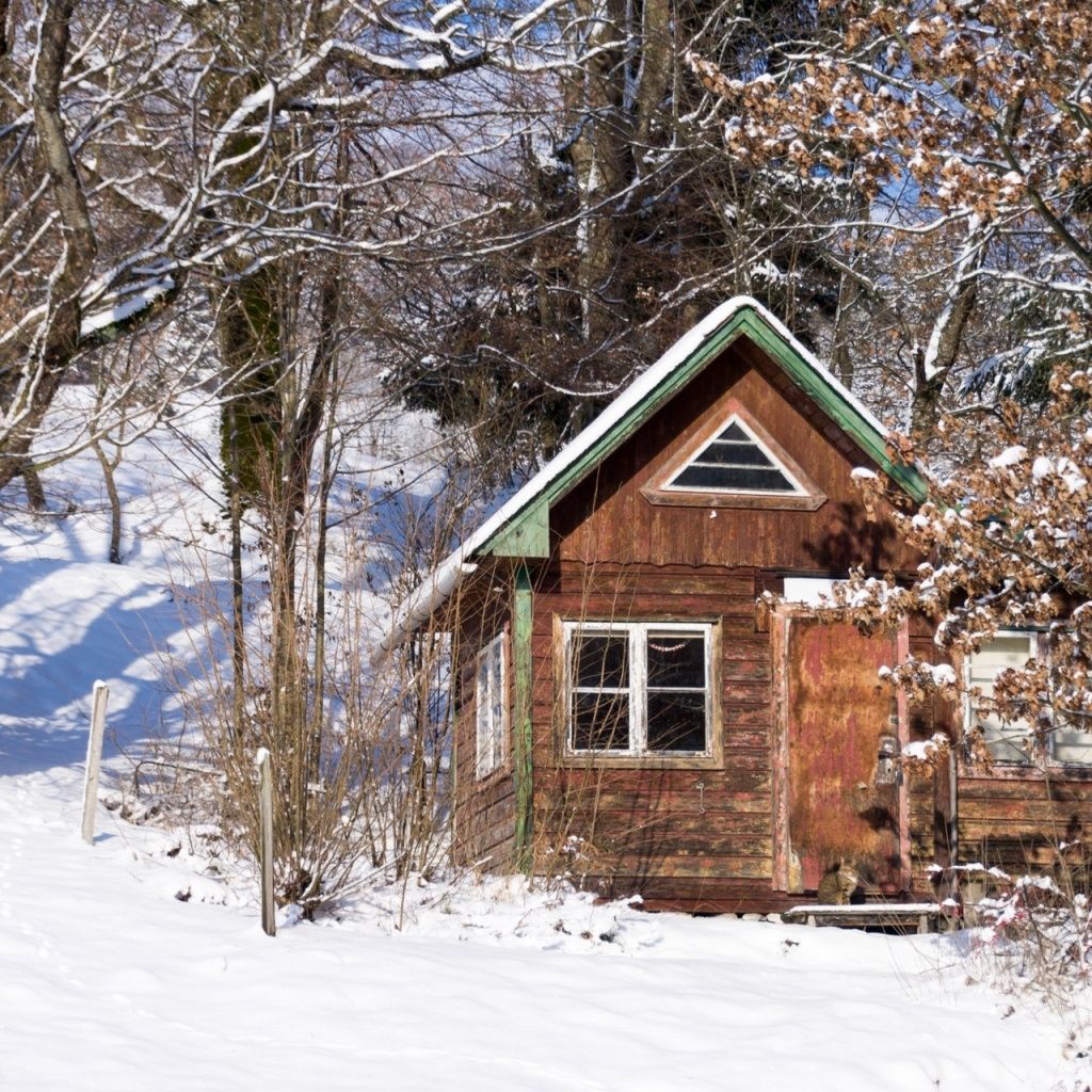 Ein kleines Holzhaus mit schneebedecktem Dach ist von schneebedeckten Bäumen umgeben. Es hat einen grünen Rand und ein kleines dreieckiges Fenster an der Vorderseite.