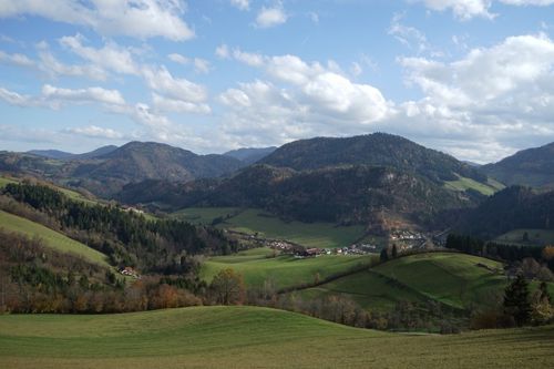 Ein landschaftlicher Blick auf ein weitläufiges, grünes Tal unter einem blauen Himmel mit Wolken, umgeben von Hügeln und Bergen, mit einem Dorf, das im Tal liegt.