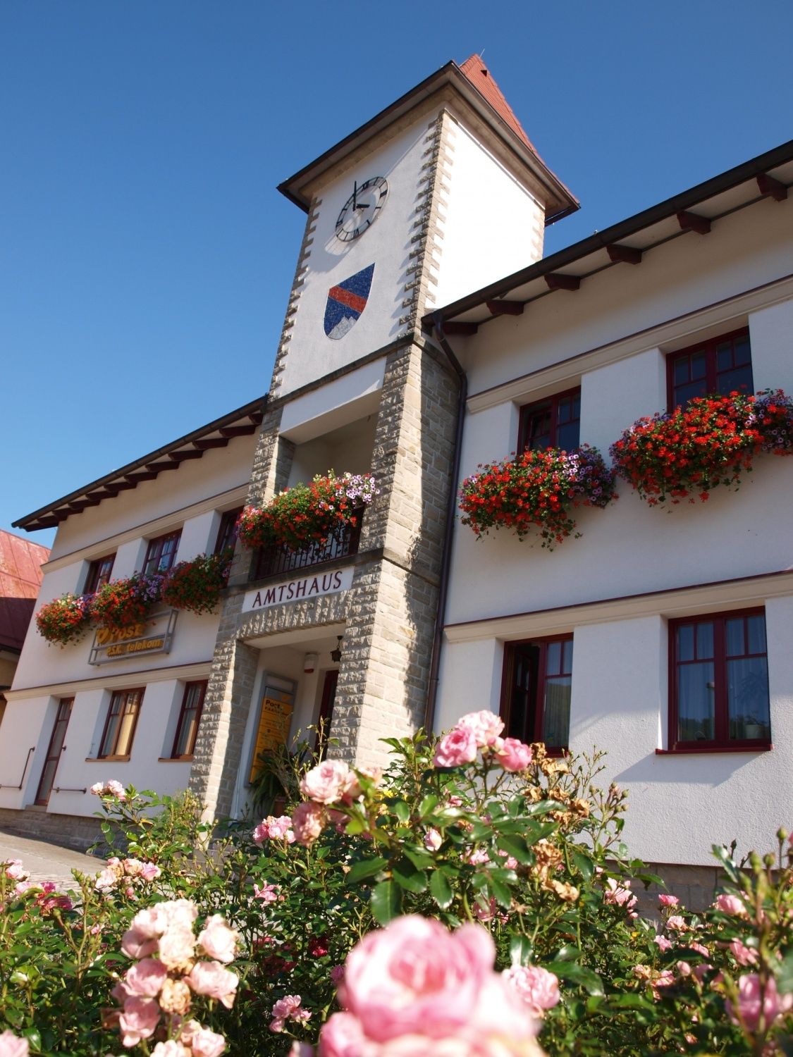 Das Amtshaus-Gebäude, geschmückt mit Blumenkästen, steht prominent mit einem Uhrenturm. Ein Schild steht Amtshaus, und rote Blumen sind an den Fenstern.