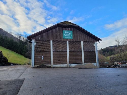 Ein braunes Gebäude mit einem grünen Schild, auf dem FWG steht. Es ist von einem grünen Feld und Bäumen umgeben, mit einem blauen Himmel und einigen Wolken.