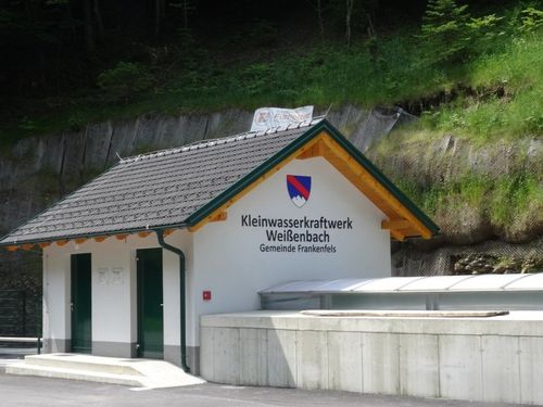 Ein weißes Gebäude mit grauem Dach ist mit 'Kleinwasserkraftwerk Weißenbach' beschriftet. Zwei Türen sind geschlossen. Ein kleiner Hügel und Bäume befinden sich dahinter.